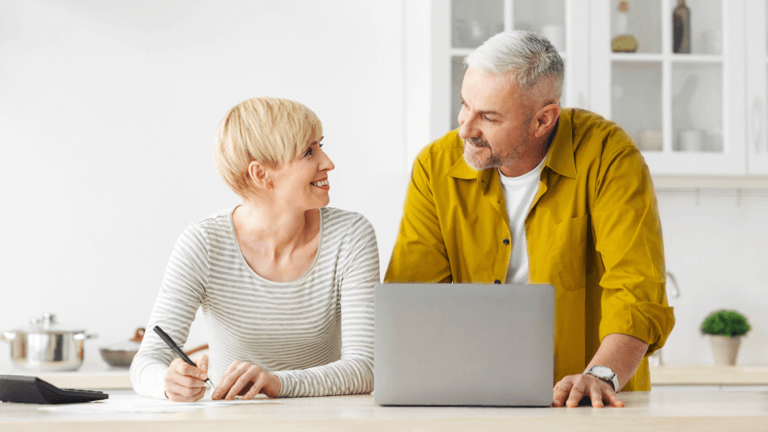 Couple at kitchen table , taking notes, and using a laptop.